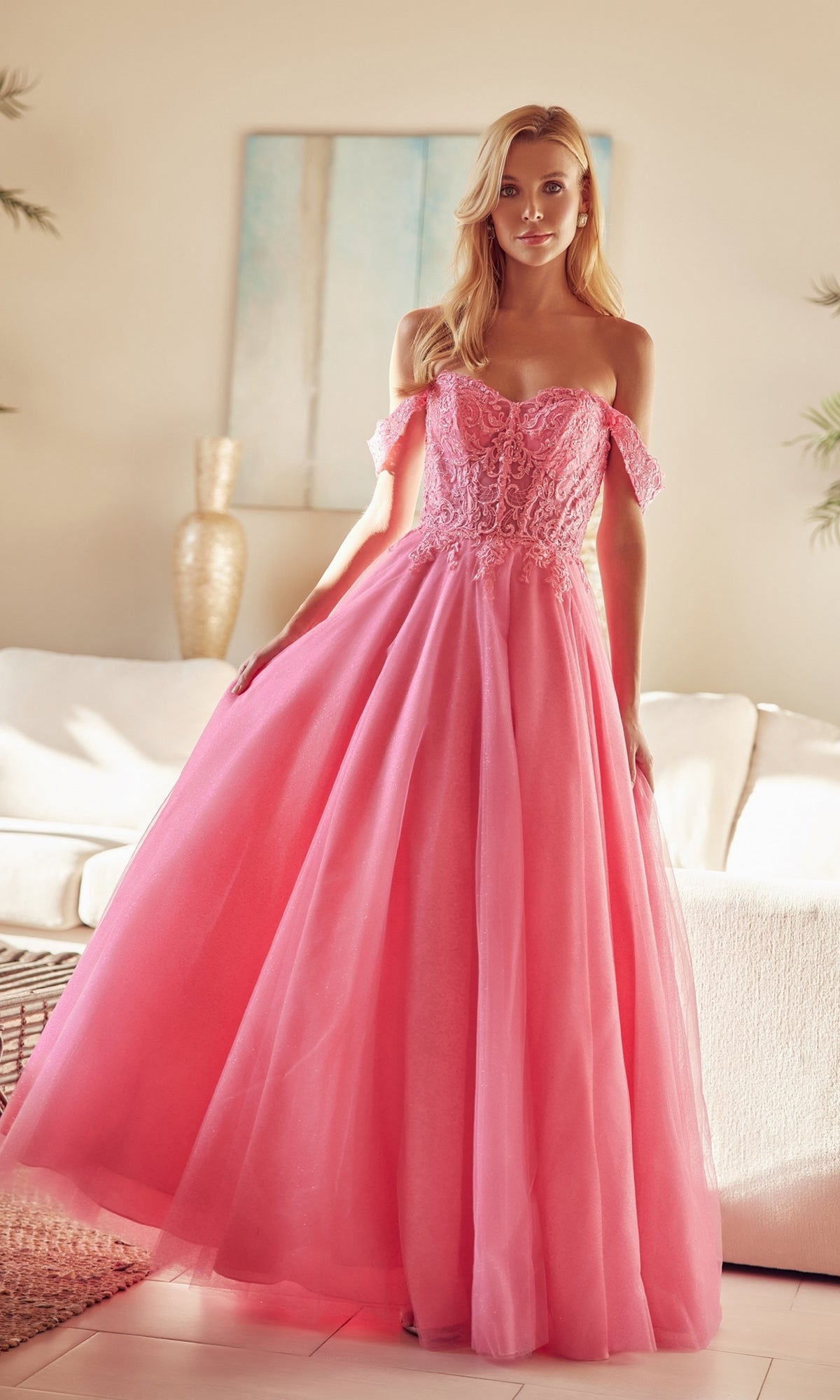 A woman stands indoors in front of a white sofa, wearing the Lace-Up Off-Shoulder Long A-Line Prom Dress 280, featuring a pink lace corset bodice and tulle details amidst neutral-toned decor.